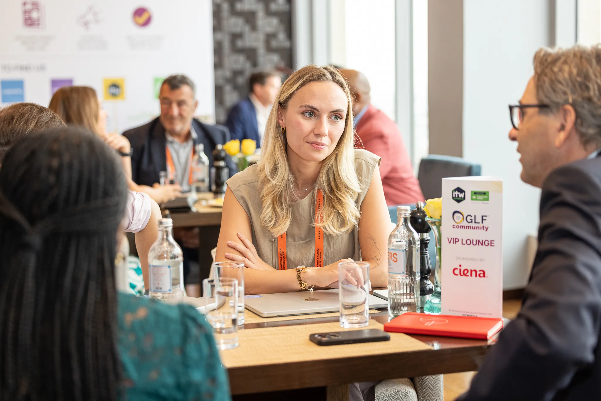A blonde woman listening attentively while sitting with other corporate delegates at a meeting table with a GLF Community VIP Lounge sign.