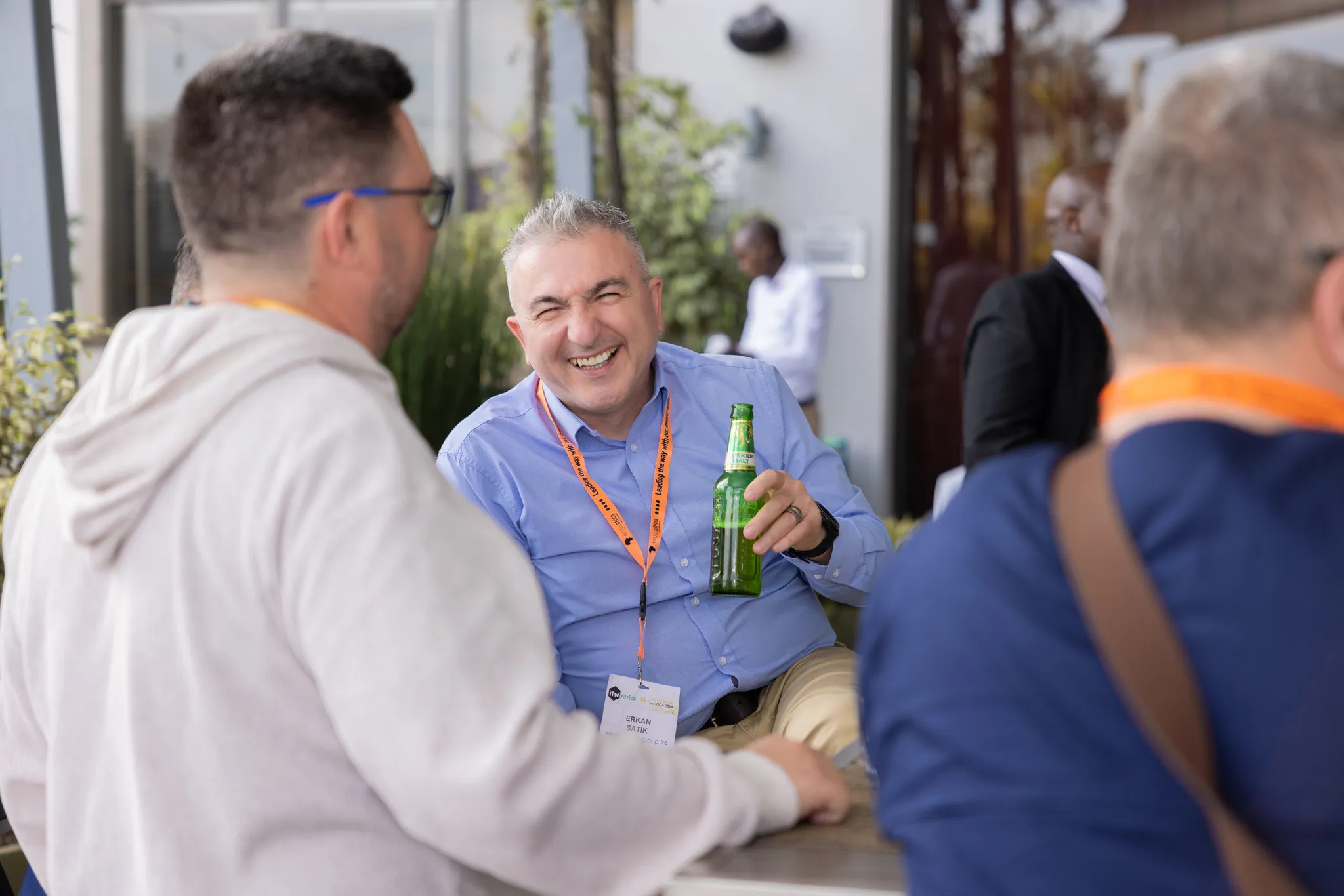 A laughing man in a light blue shirt holding a green bottle of beer and chatting with colleagues outdoors during a corporate happy hour event.