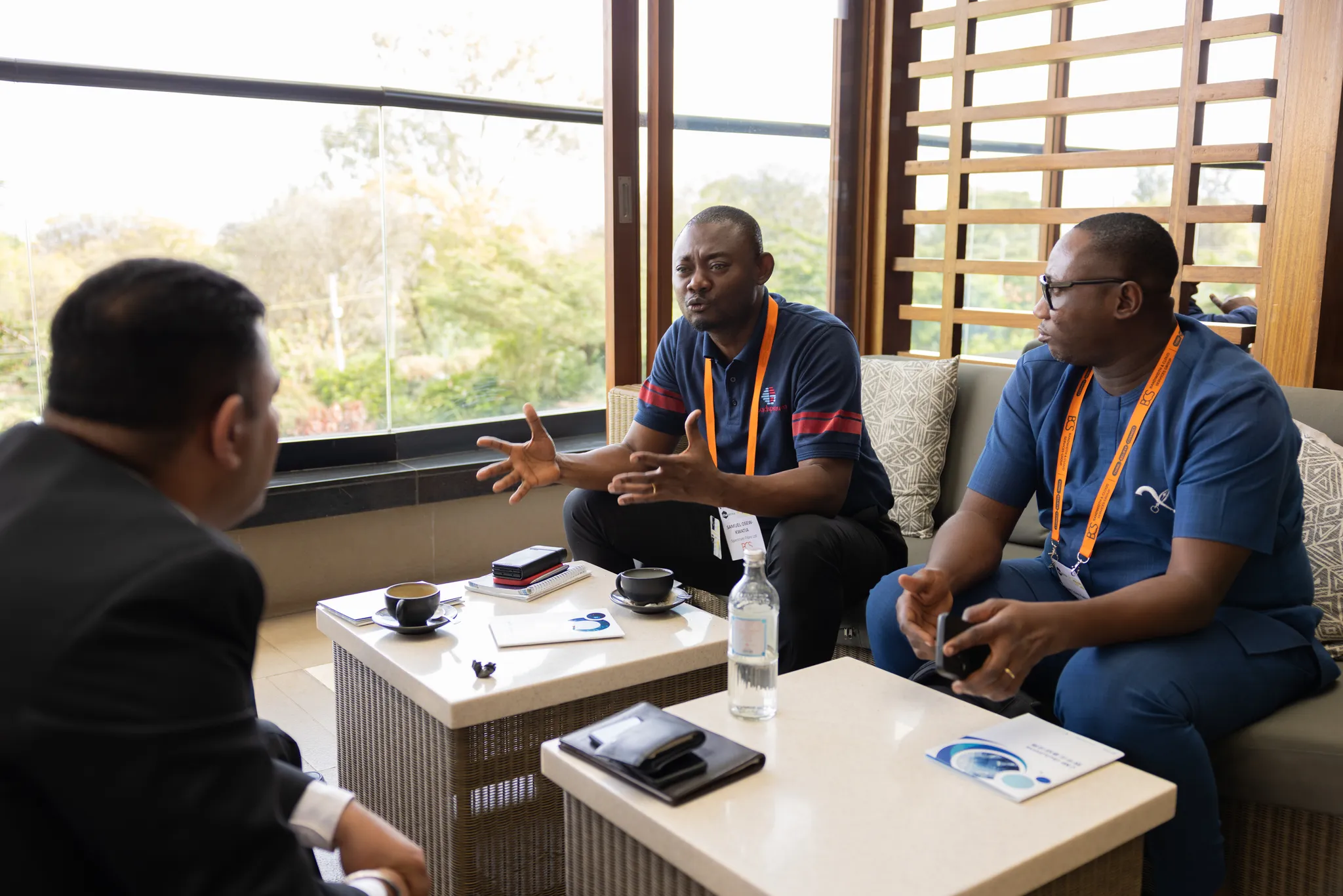 A group of three male event attendees sitting on comfortable outdoor lounge chairs, gesturing and discussing business over coffee and water.