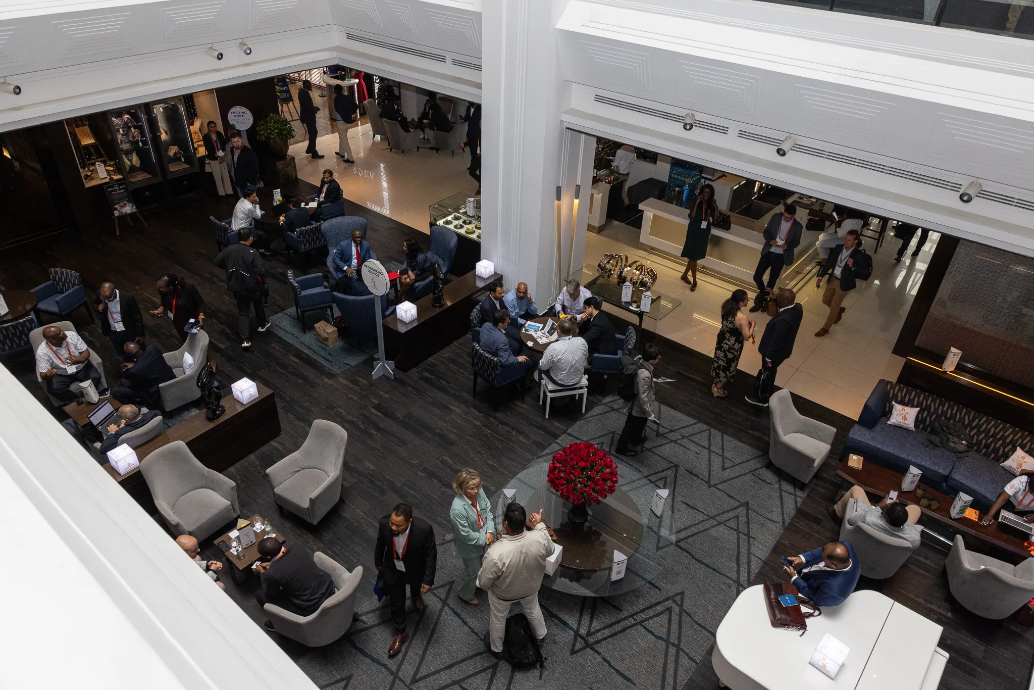 A high-angle view looking down into a large, modern hotel lobby arranged into multiple lounge clusters where conference delegates gather to chat.