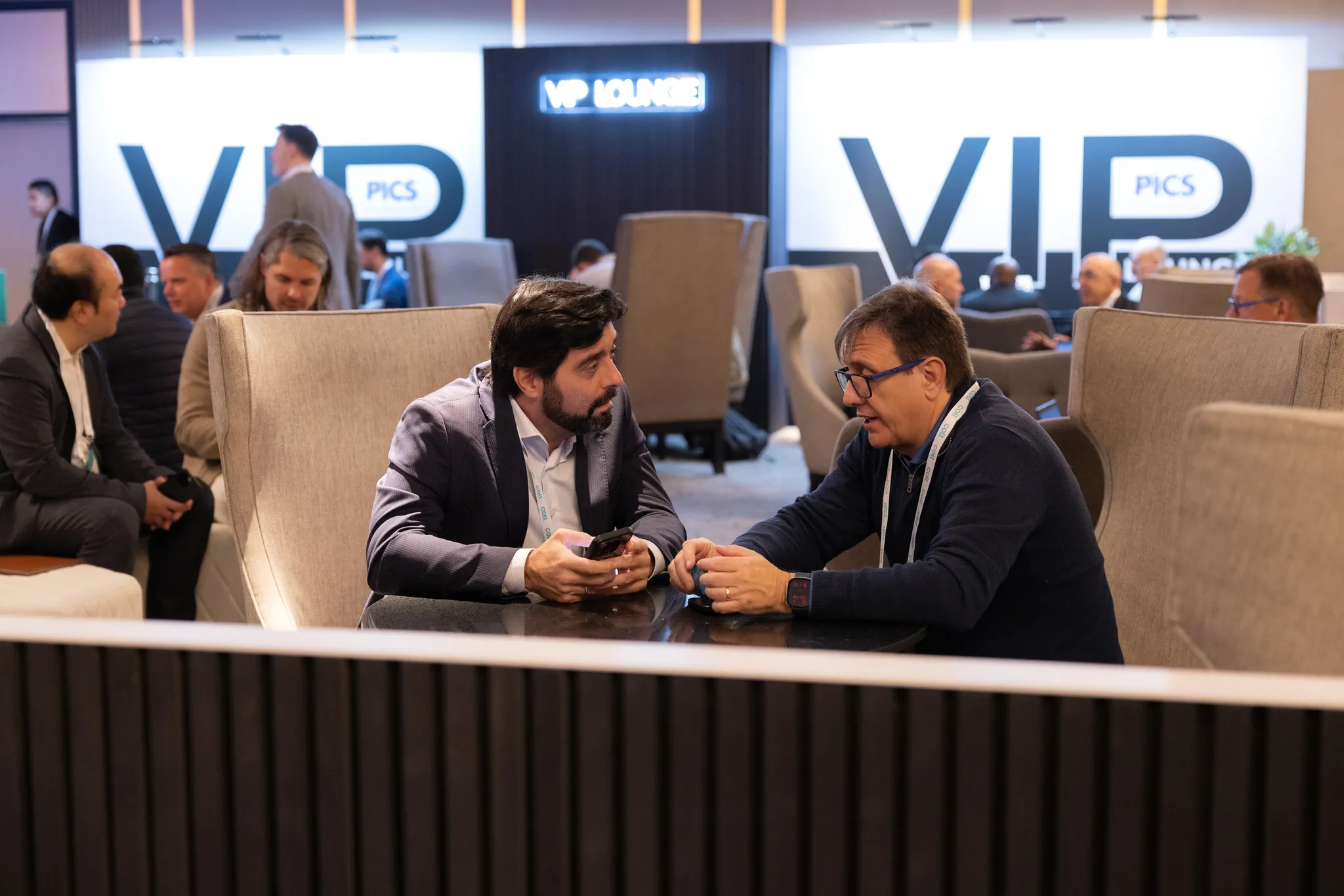 Two men in professional attire conversing across a table in high-backed armchairs within a conference VIP lounge space.