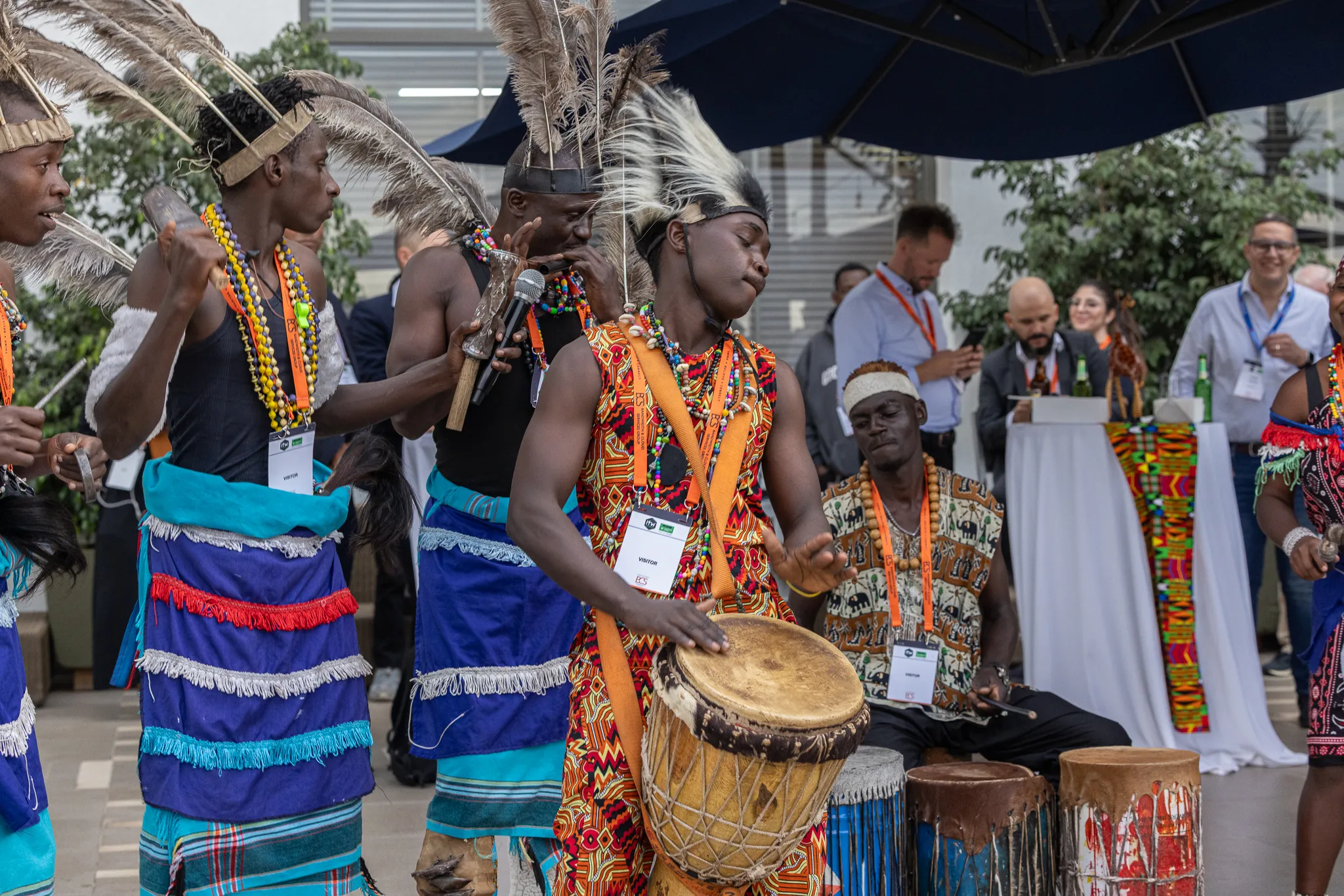 Traditional African dancers and percussionists in colorful cultural attire performing with drums at an outdoor corporate drinks reception while guests socialize in the background.