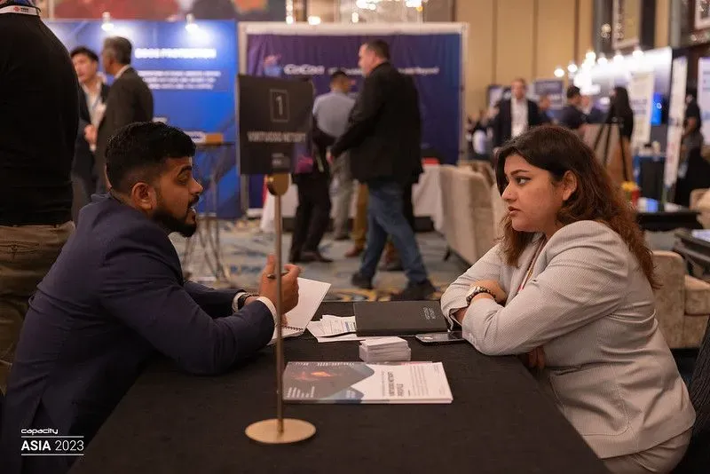 Two attendees engaged in a focused one-to-one business meeting at a table during the Capacity Asia 2023 event. The background shows a busy networking hall with various exhibition booths and industry professionals.
