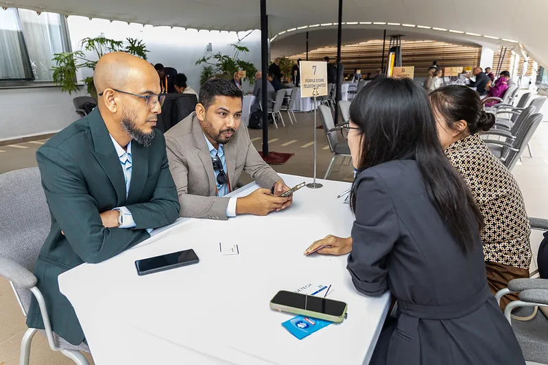 Four conference attendees are seated around a meeting table in a networking area, discussing business opportunities during a hosted buyer-style meeting session. Meeting table signage is visible, with additional delegates and networking tables in the background of the event venue.