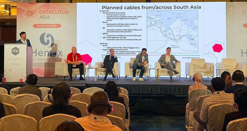 A photograph of a panel discussion taking place on stage at the Datacloud Asia conference. One speaker stands at a podium on the far left, while four other panelists are seated in white chairs across the stage. Behind the panelists, a large presentation screen displays a slide titled 