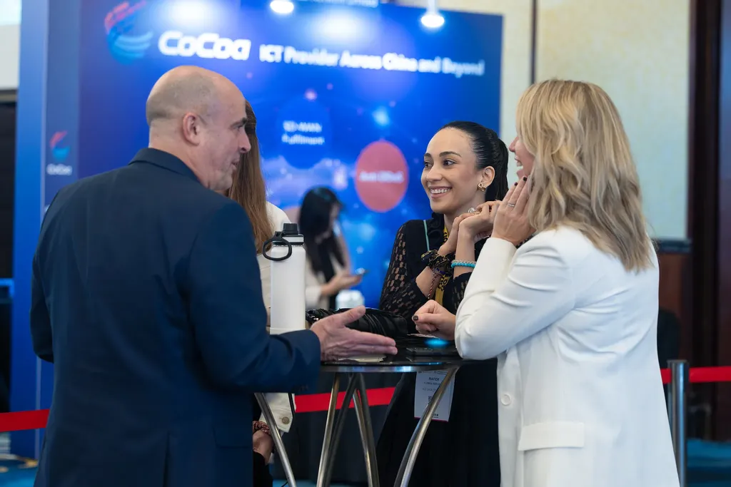 Three professionals networking around a high table at a Datacloud Asia event. A man in a blue suit talks with two women, one in a white blazer and one in a black lace top, in front of a blue exhibition backdrop for CoCoa, an ICT provider.