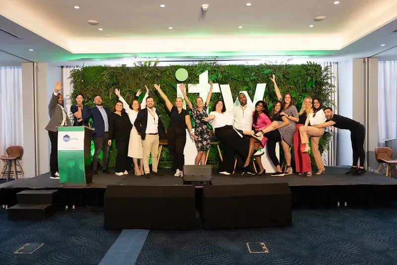 A group of diverse event professionals posing playfully on a stage in front of a large green plant wall featuring the white 