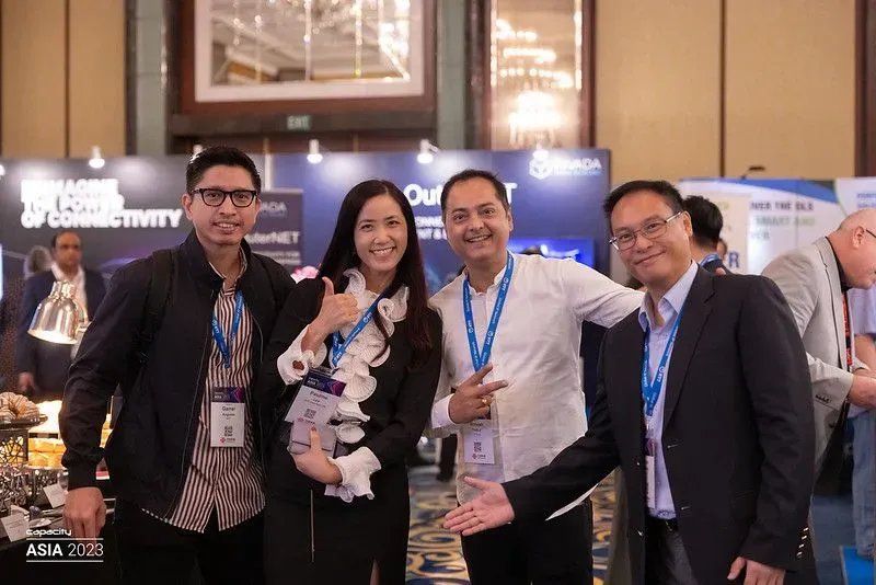 Four professionals smiling and posing together at the Datacloud Asia 2023 event. The group includes three men and one woman, all wearing event lanyards, in a brightly lit conference hall with exhibition booths and 