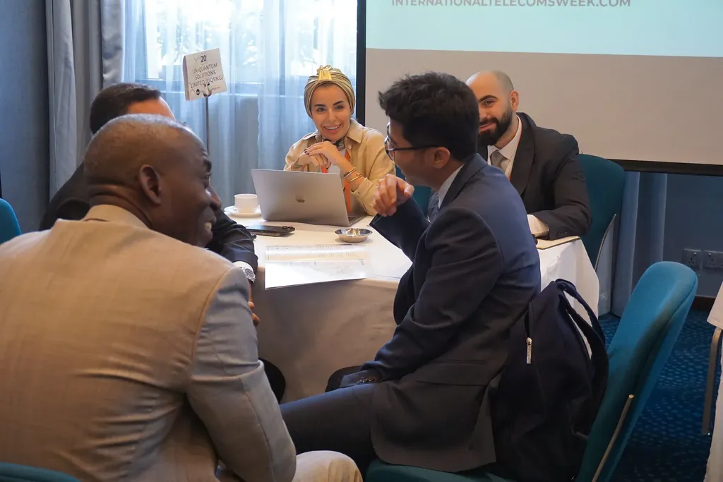 A group of diverse professionals in business attire engaged in a networking discussion around a table at ITW Asia, with laptops and notebooks present.