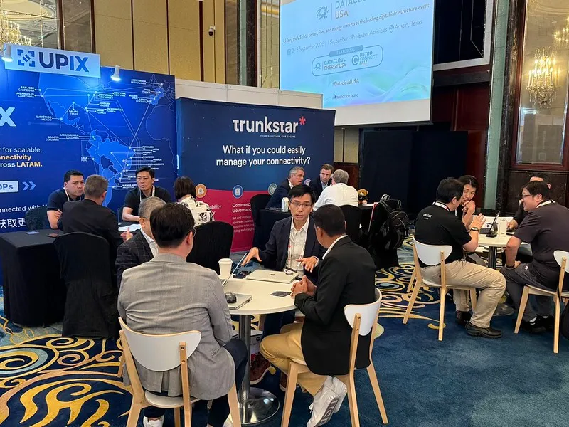Attendees take part in hosted networking meetings at a business conference, seated at small tables in an exhibition hall with exhibitor stands and event branding visible in the background.