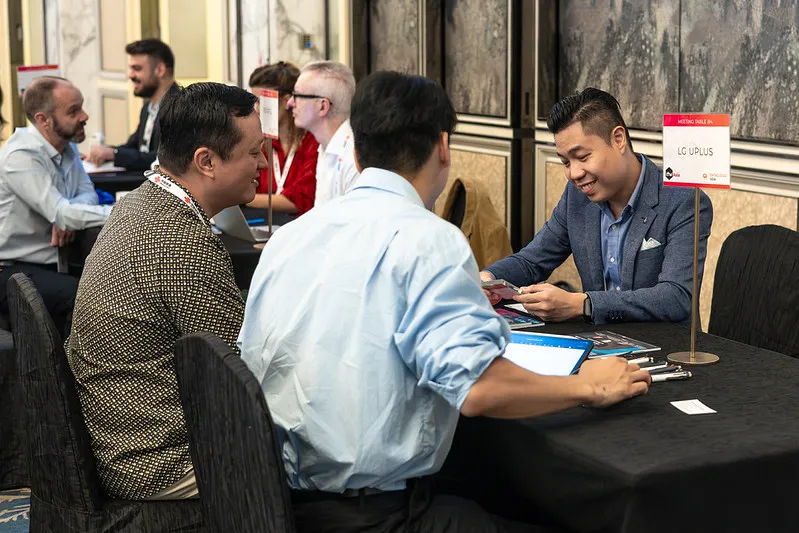 Several attendees are seated at a branded networking table in a conference meeting area, holding a business discussion during a one-to-one networking session. Additional meeting tables and delegates are visible in the background, highlighting the organised networking environment of the event.