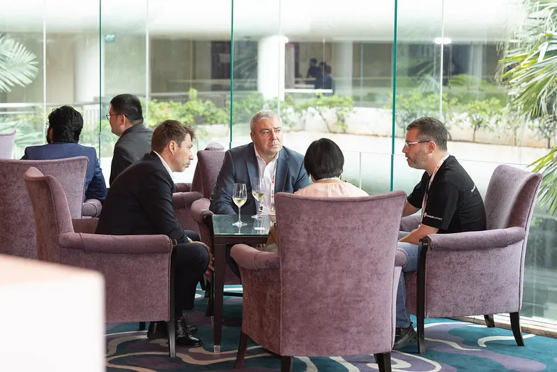 Conference attendees seated in a lounge area take part in a business networking discussion around a small table, with floor-to-ceiling windows and a bright event venue visible in the background.