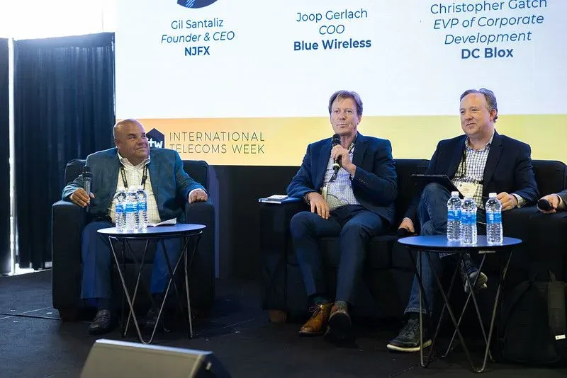 A panel of three industry executives speaking at an International Telecoms Week (ITW) event. The speakers, including leadership from NJFX, Blue Wireless, and DC Blox, are seated on a stage with a digital backdrop displaying their names and titles.