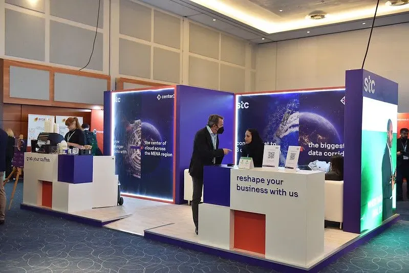 A modern purple and white exhibition booth for stc at a Datacloud Asia event. Two professionals engage in a business discussion near a desk featuring the slogan 