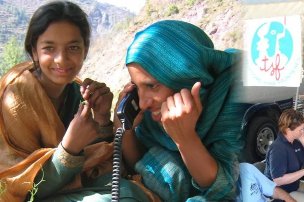 A woman wearing a teal headscarf smiles while using a satellite phone in a mountainous outdoor region. Next to her, a younger person in a green and gold shawl looks on. The image includes a 