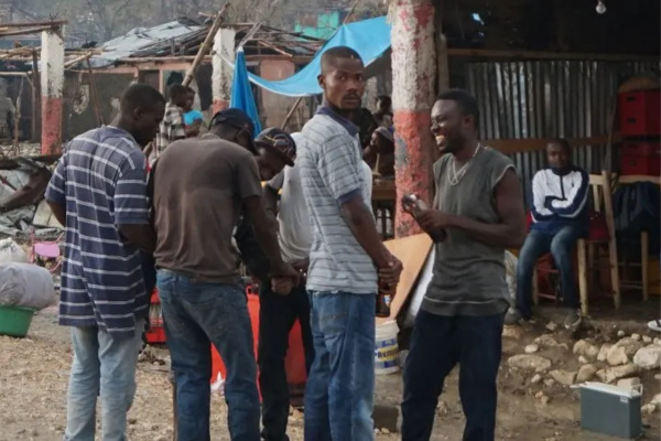 A group of men gathered in a rural outdoor setting. One man in a grey tank top is laughing while holding a small device, while others stand nearby in conversation. The background features modest structures with corrugated metal roofing and a blue flag.