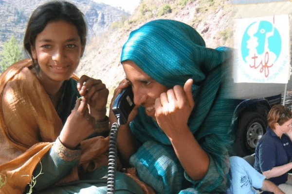 A woman wearing a teal headscarf smiles while using a satellite phone in a mountainous outdoor region. Next to her, a younger person in a green and gold shawl looks on. The image includes a 