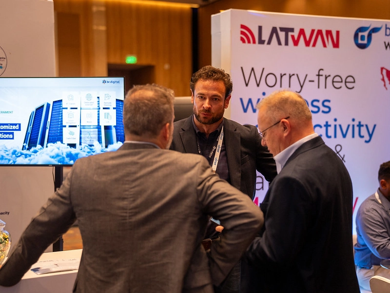 Three event attendees networking near exhibitor booths featuring br.digital and LATWAN branding at a telecommunications trade show.