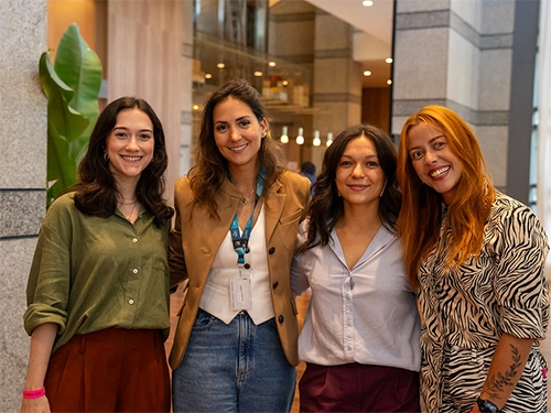 Four women posing side-by-side and smiling in a brightly lit modern venue atrium. The woman second from the left wears a tan blazer over a white top with an event lanyard and badge, while the woman on the far right wears a black and white zebra-patterned outfit.