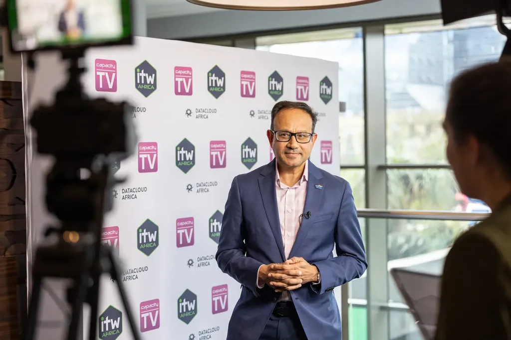 Behind-the-scenes shot of a man wearing glasses, a pink shirt, and a navy blue suit jacket standing in front of a branded media backdrop (featuring Capacity TV, ITW Africa, and Datacloud Africa logos). A camera on a tripod is silhouetted in the foreground on the left, and an interviewer's shoulder is visible on the right.