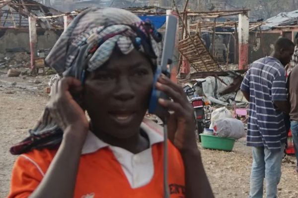 A street scene in a developing area or disaster zone, depicting a community gathering around communication services. In the close-up foreground, a person wearing an orange shirt and a patterned head wrap speaks on a large satellite phone. In the background, multiple local men stand or sit near basic structures and shelters with damaged roofs, some looking toward the camera or talking amongst themselves.
