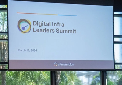 A photo of a large projection screen inside a brightly lit venue with outdoor foliage visible in the background. The slide shows the logo and text 