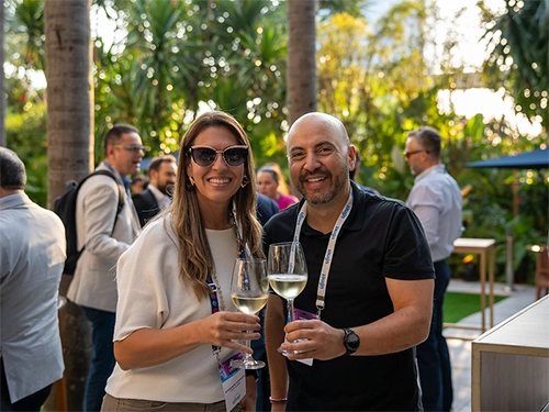 A smiling woman wearing cat-eye sunglasses and a white top stands alongside a smiling, bald man in a black polo shirt, both holding glasses of white wine at an outdoor networking reception surrounded by palm trees.