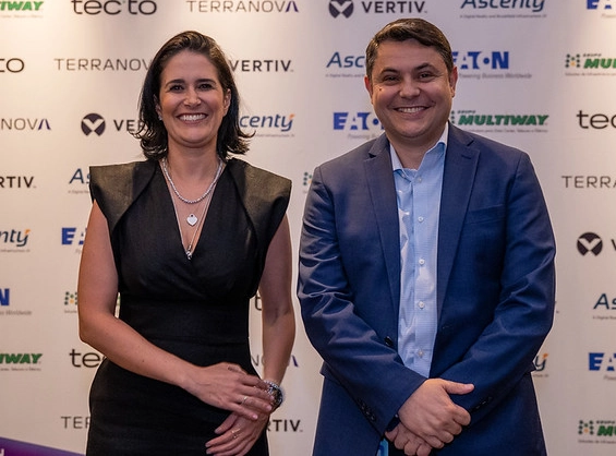 A smiling woman in a sleeveless black dress and a smiling man in a blue suit pose side by side in front of a sponsor backdrop wall featuring logos such as Vertiv, Ascenty, Eaton, and Terranova.