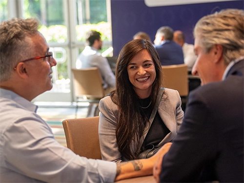 A smiling woman with long brown hair sits at a conference table, engaged in conversation with two men whose backs/profiles face the camera.
