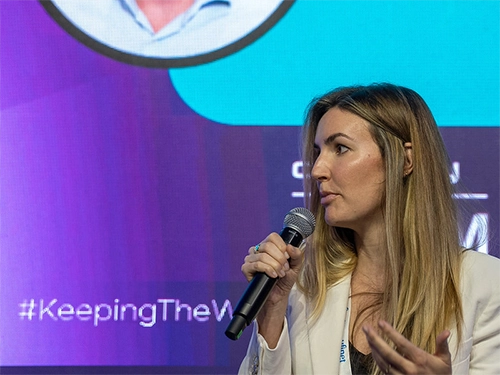 A blonde woman in a white blazer speaking into a microphone with hand gestures on stage, in front of a purple and teal backdrop with the hashtag 