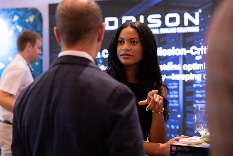 An engaged professional woman gestures toward a digital infrastructure cooling solutions display while speaking with an attendee at a Datacloud USA exhibition booth.