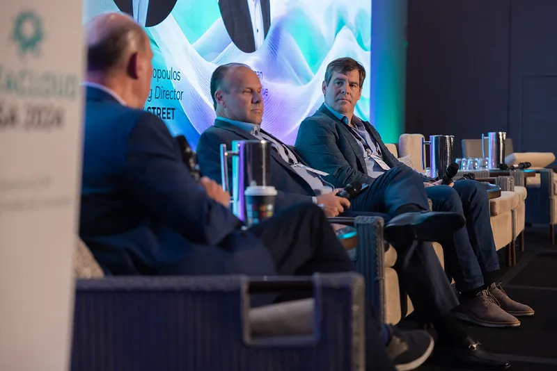Three men sitting on a stage, talking. The gentlemen with his back towards the camera is holding a microphone. All three are dressed in suits. 