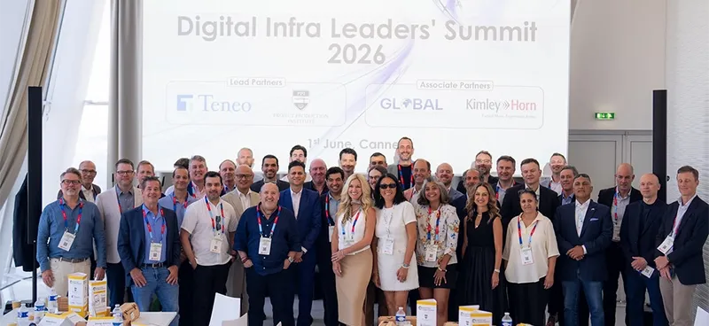 A group of business professionals smiling and posing together at the Digital Infra Leaders' Summit 2026 in Cannes, standing before a large projection screen displaying event partner logos including Teneo, PPI, Global Commissioning, and Kimley-Horn.