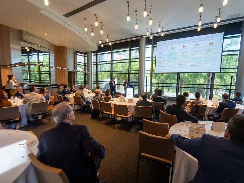 A conference room filled with professionals seated around round banquet tables, listening to speakers standing in front of floor-to-ceiling windows next to a large digital display showing a Latin American market analysis presentation.