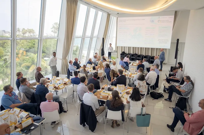 An overhead view of a brightly lit seminar hall overlooking the coast through large floor-to-ceiling windows, where attendees sit at tables facing a speaker standing beside a presentation screen titled 
