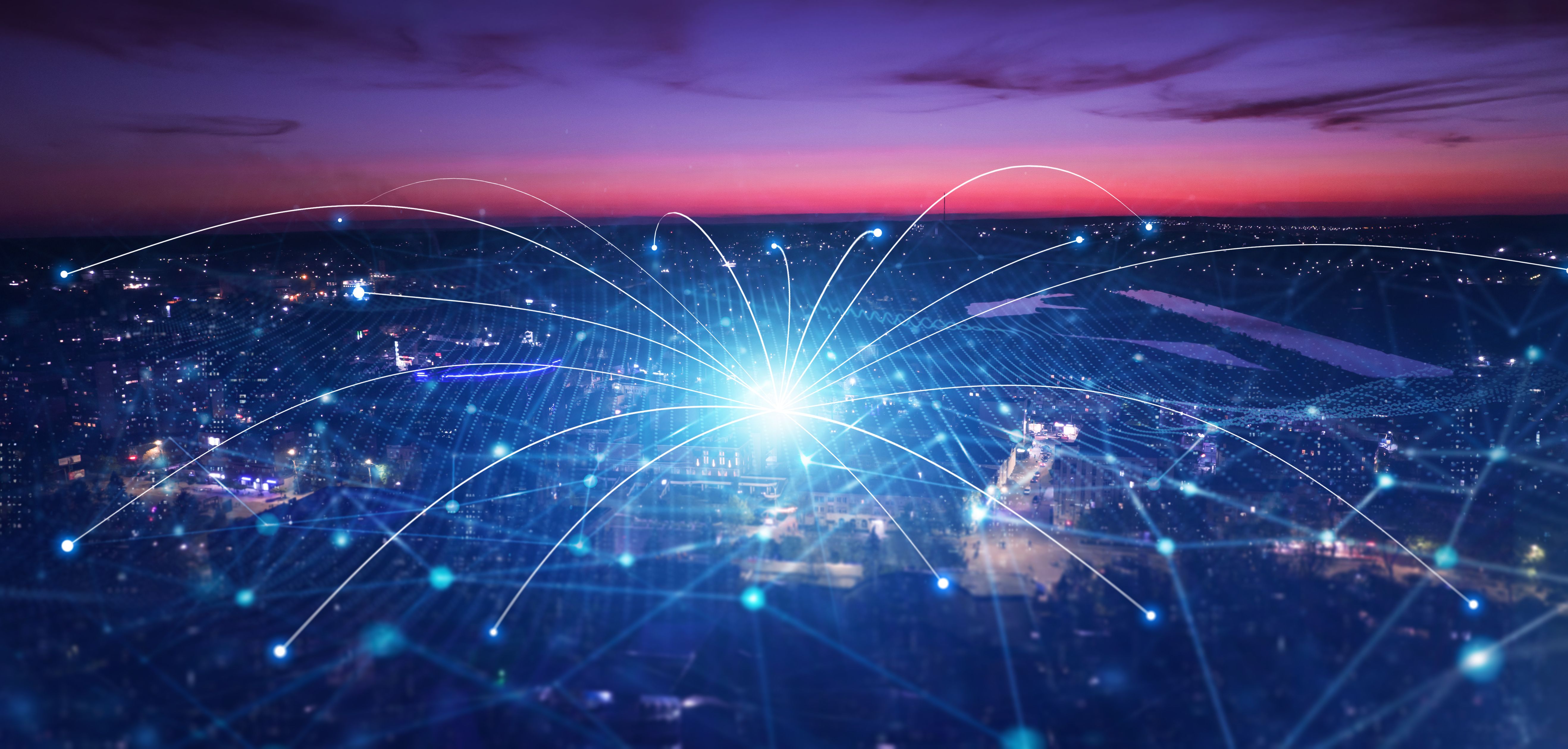 A wide-angle aerial view of a city at twilight with a vibrant purple and orange sky. A glowing blue digital network overlay originates from a central bright light point, with data lines and nodes curving across the cityscape to symbolise connectivity.