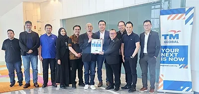 A group of nine people in business-casual and formal attire standing in a modern office lobby. In the center, two representatives hold up an official framed Tier III data center certification plaque. To their right stands a blue and white TM Global pull-up banner displaying the slogan "YOUR NEXT IS NOW."