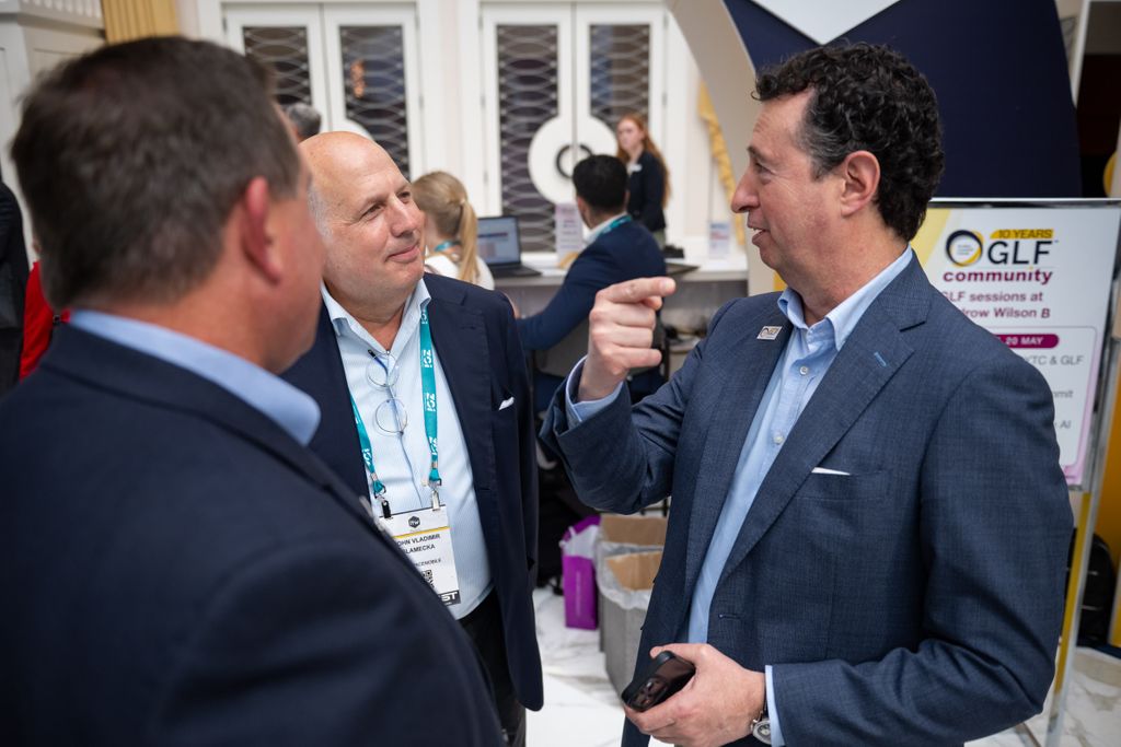 A professional networking interaction at an exhibition. A woman in a sharp dark blue suit is smiling warmly while shaking hands with a man wearing a light patterned shirt, glasses, and a backpack. In the background, a lime green display wall features corporate information, text regarding wholesale services, and a QR code.