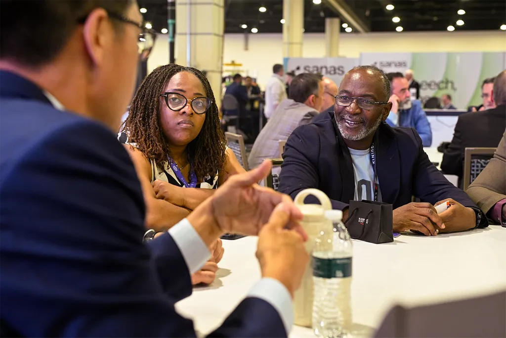 An over-the-shoulder shot of a business meeting taking place at a round table inside a large convention or event hall. In the foreground, a man in a blue suit is seen from behind, gesturing with his hands as he speaks. Across the table, a woman with braided hair and glasses listens intently with her arms crossed, while a man next to her with glasses and a grey beard smiles warmly at the speaker. Other attendees and event booths, including one with the text 