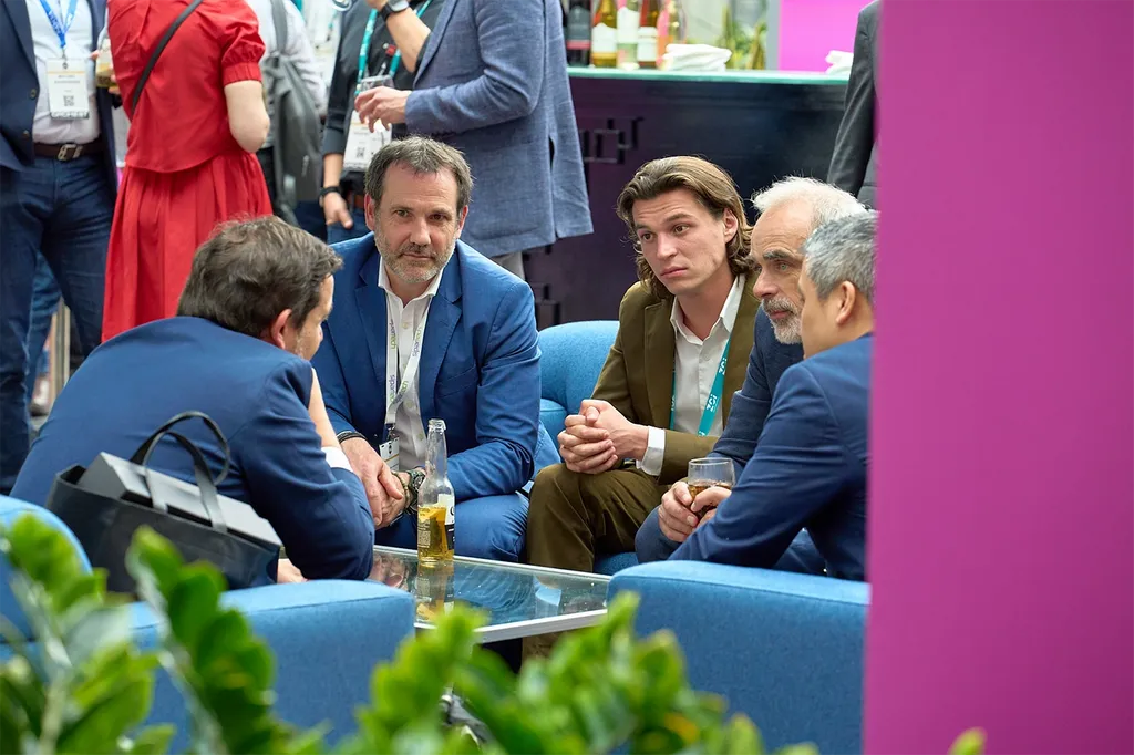 A group of business professionals sitting in blue armchairs around a small glass coffee table, deeply engaged in conversation at a networking event. A man in a blue suit looks on attentively from the left side of the circle, while a younger man in an olive-green suit and an older man with gray hair listen closely on the right. In the foreground, a prominent pink structural pillar stands on the right, and green plant foliage frames the bottom-left corner. Additional event attendees are visible talking in the blurred background.