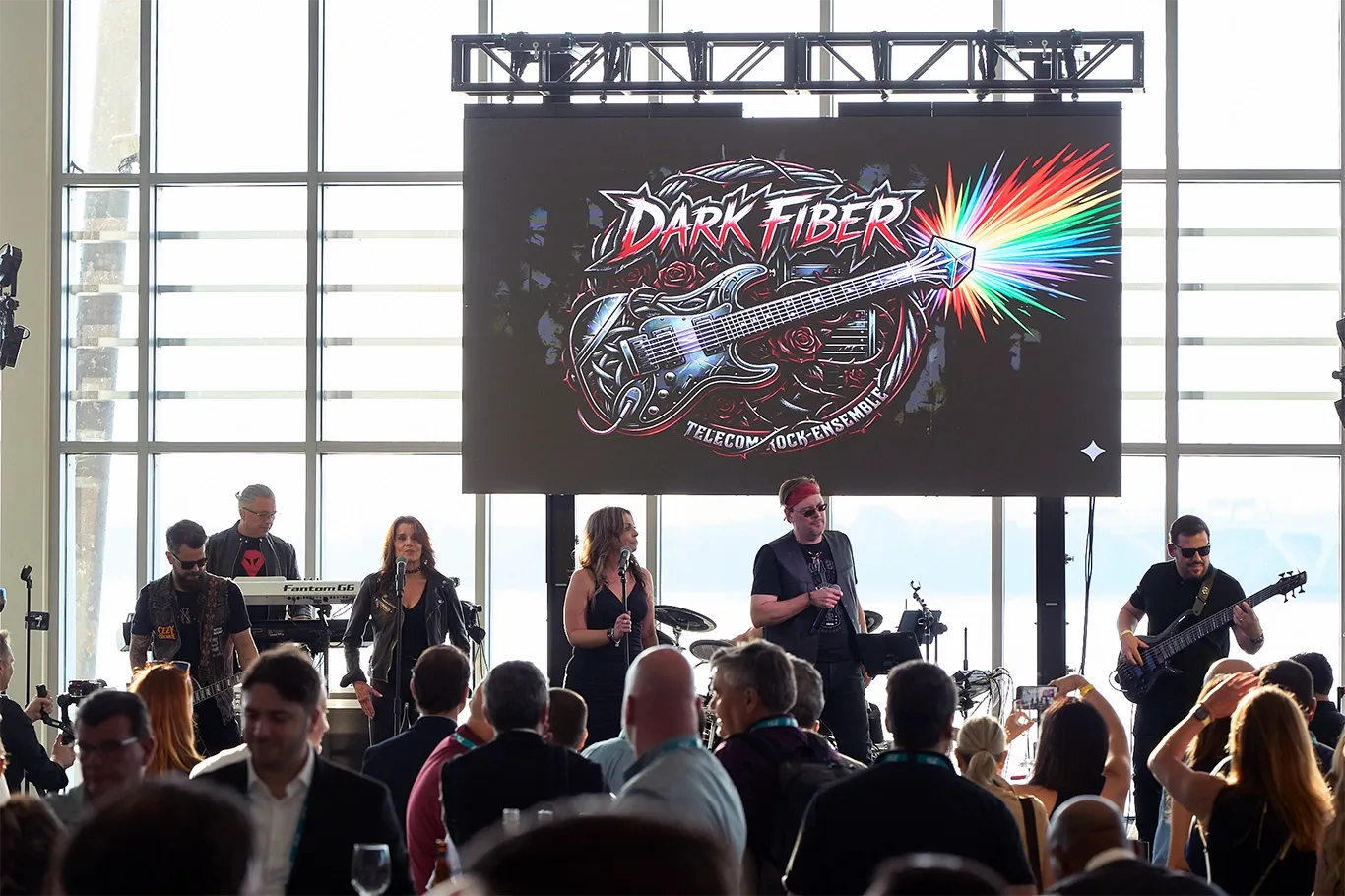 A live musical performance taking place on a stage in front of a seated audience. On stage, a rock band consisting of multiple vocalists, guitarists, and a keyboard player is performing. Hanging directly above them is a large digital screen displaying a stylized logo with an electric guitar and roses that reads 