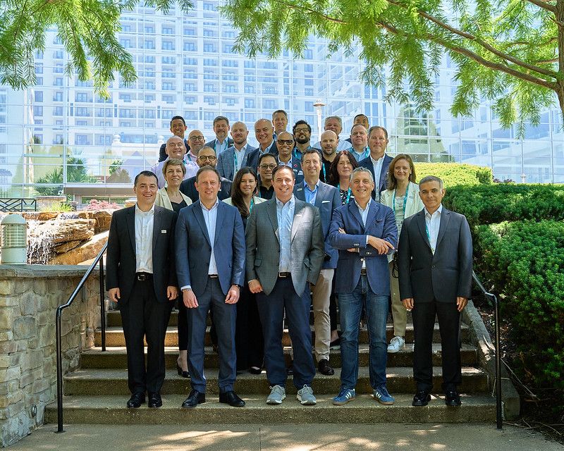 A group portrait of about twenty professionals, mostly men and a few women, smiling and posing together on outdoor stone steps. They are dressed in business casual and formal attire. The background features a large, modern glass building, lush green trees framing the top, and a small rock waterfall feature to the left.