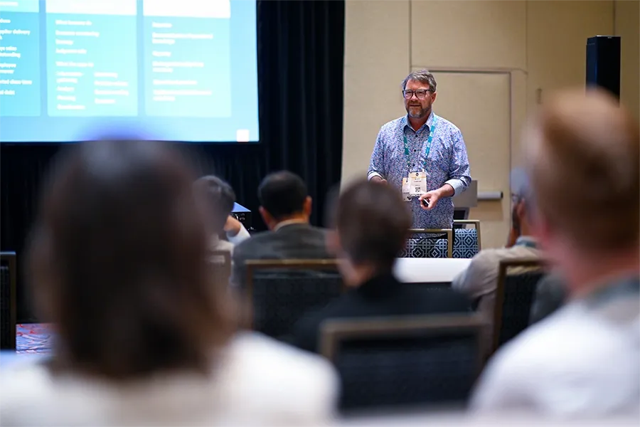 A presenter standing at the front of a conference room, speaking to an audience seated in rows. The presenter is a man with glasses and a beard, wearing a patterned blue button-down shirt and an event lanyard. To his left, a large presentation screen displays structured text blocks and columns. The audience members in the foreground are captured from behind and are softly out of focus, drawing attention to the speaker.