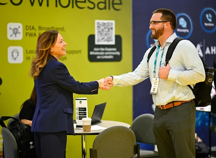 A side-profile, medium shot of a positive interaction at a professional event. On the left, a smiling woman with wavy brown hair wearing a dark blue blazer shakes hands with a man on the right. The man, wearing glasses, a beard, a light-patterned button-down shirt, a brown belt, and grey trousers, smiles warmly while carrying a black backpack over his shoulders. In the background, a vibrant chartreuse wall features a QR code and partial text reading 