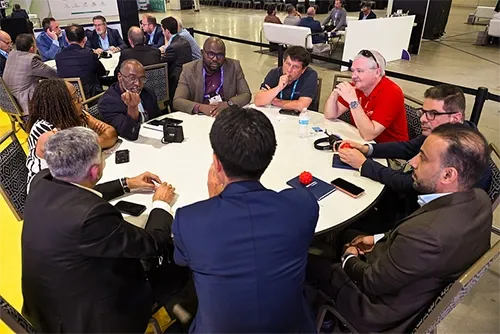 A high-angle shot looking down at approximately ten diverse professionals seated in a circle around a large, white round table in an expansive exhibition hall. The attendees are dressed in business casual attire and appear highly engaged, with several looking toward a man on the left who is speaking and gesturing. On the table, various notebooks, smartphones, water bottles, and small red stress balls are scattered. The background shows a spacious, open conference floor with other tables and seating zones visible in the distance.