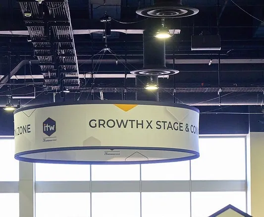 A medium shot looking upward at a large, cylindrical sign suspended from an open industrial ceiling. The white circular banner features a dark blue border and displays the 