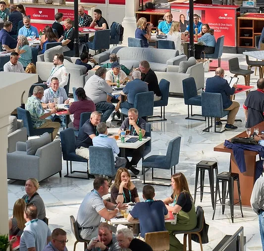 A wide, high-angle shot looking down at a bustling conference networking lounge situated in a spacious indoor atrium with a polished white marble floor. Dozens of attendees, many wearing event lanyards, are seated in small groups across a mix of grey modern sofas, armchairs, and small square tables with dark blue upholstered chairs. In the background, red exhibition booths with 