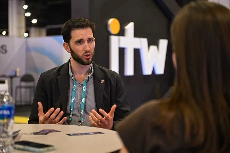 A medium, over-the-shoulder shot of a man with a dark beard and short hair speaking dynamically. He is wearing a grey button-down shirt under a black blazer, along with a teal conference lanyard. The back of a woman's head with dark hair is visible in the blurred foreground on the right. In the background, a large dark grey structure displays a white 