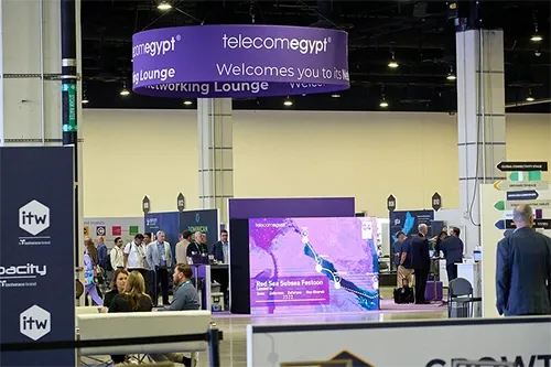 A wide, eye-level shot of a conference exhibition hall. Suspended from the high ceiling in the center is a large, cylindrical purple sign reading 