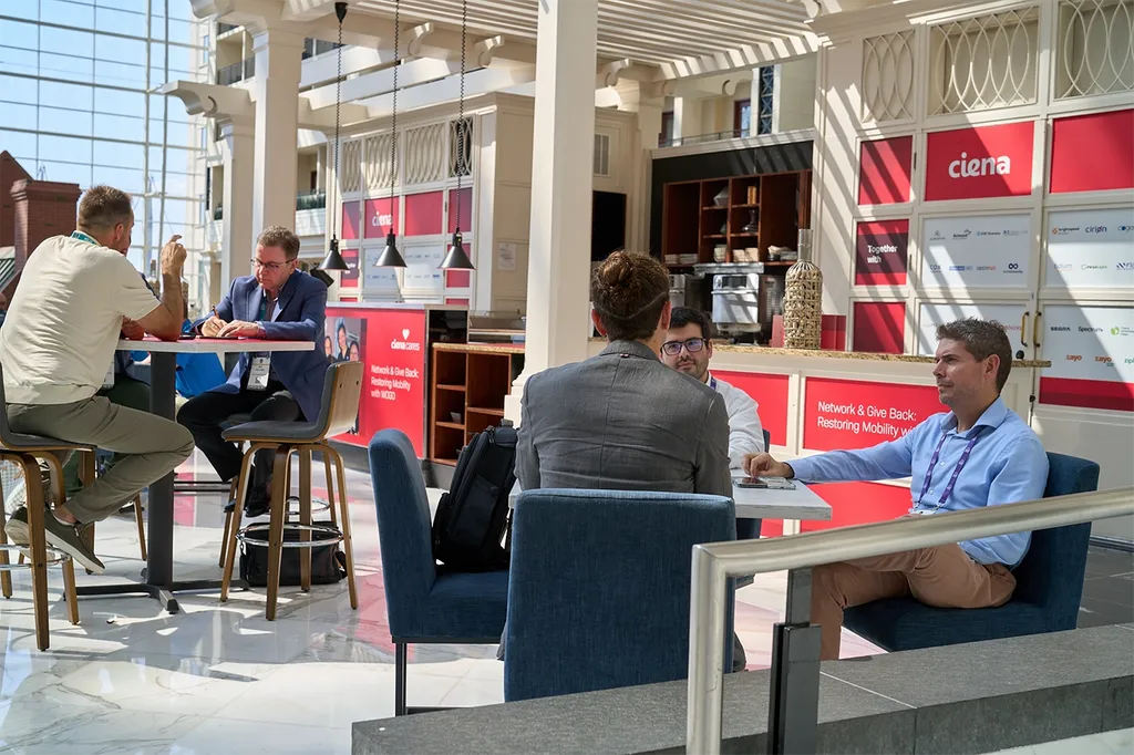 A bright, sunlit indoor atrium configured for business meetings. In the foreground, men are seated at a low table in blue armchairs, engaged in discussion. In the background, other professionals sit at a high-top table on barstools, writing and talking. The surrounding structures are decorated with corporate branding, including prominent red panels for 